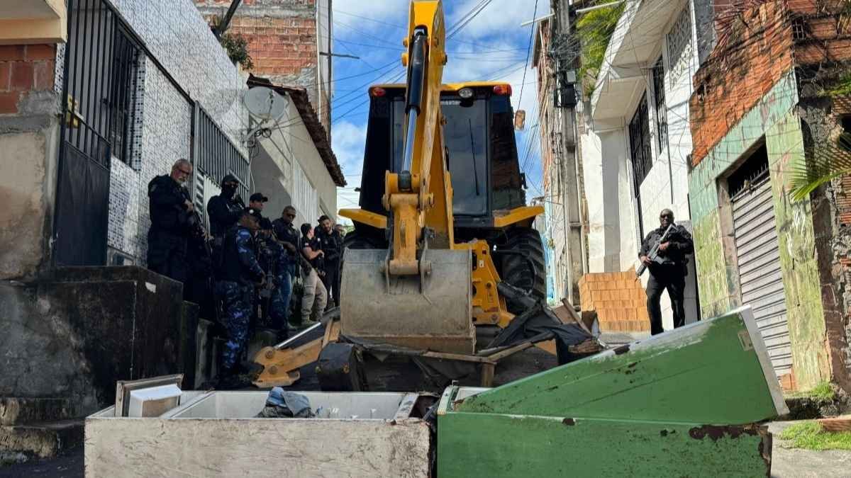 **Texto alternativo resumido da imagem:** Uma retroescavadeira remove barricadas feitas com geladeiras e entulhos em uma rua estreita de Salvador, enquanto vários policiais armados observam e dão apoio à operação.