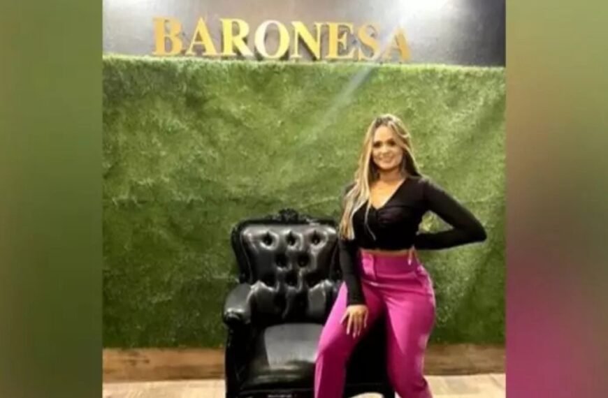 Justiça solta cunhada de Marcola -Mulher em pé ao lado de uma poltrona preta, posando para foto em frente a uma parede verde com a palavra “BARONESA” escrita em letras douradas no topo. Ela veste blusa preta de manga longa e calça rosa.