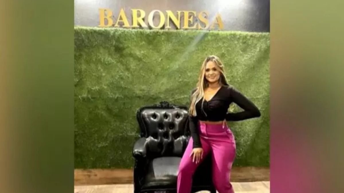 Justiça solta cunhada de Marcola -Mulher em pé ao lado de uma poltrona preta, posando para foto em frente a uma parede verde com a palavra “BARONESA” escrita em letras douradas no topo. Ela veste blusa preta de manga longa e calça rosa.