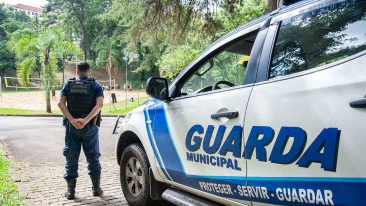 Um guarda municipal de uniforme azul, de costas, está em pé próximo a uma viatura da Guarda Municipal branca com faixas azuis. Ao fundo, há uma quadra de areia com pessoas jogando vôlei, em um ambiente arborizado.