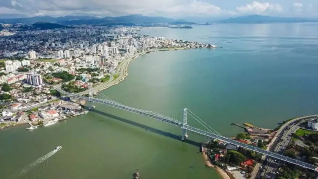 A imagem mostra uma vista aérea de Florianópolis, destacando a Ponte Hercílio Luz ligando a ilha ao continente, com a cidade, prédios e a orla ao fundo.