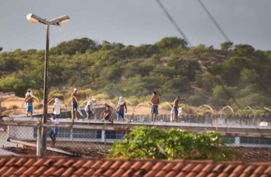 O Massacre de Alcaçuz: Grupo de presos sobre o telhado de um presídio durante rebelião, alguns de torso nu e com armas improvisadas nas mãos, cercados por arame farpado; ao fundo, vegetação em área de morro