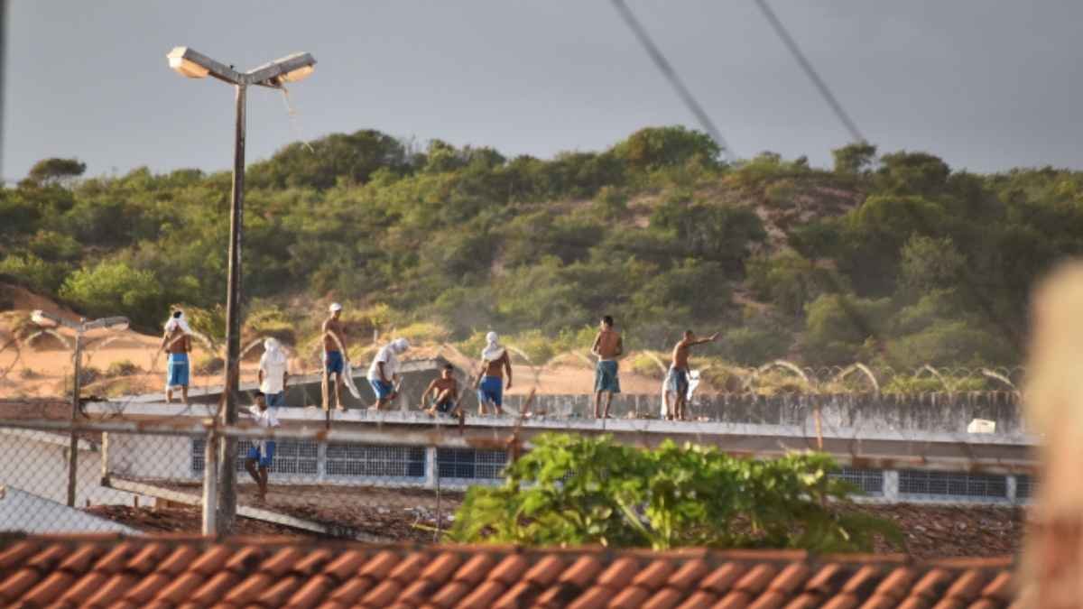 O Massacre de Alcaçuz: Grupo de presos sobre o telhado de um presídio durante rebelião, alguns de torso nu e com armas improvisadas nas mãos, cercados por arame farpado; ao fundo, vegetação em área de morro