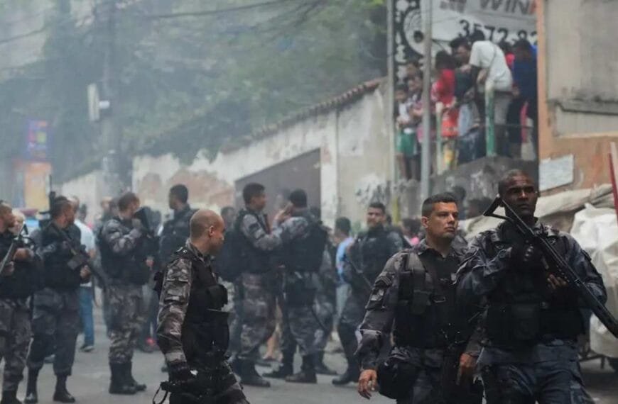 "Policiais fortemente armados em operação em comunidade, enquanto moradores observam das casas.