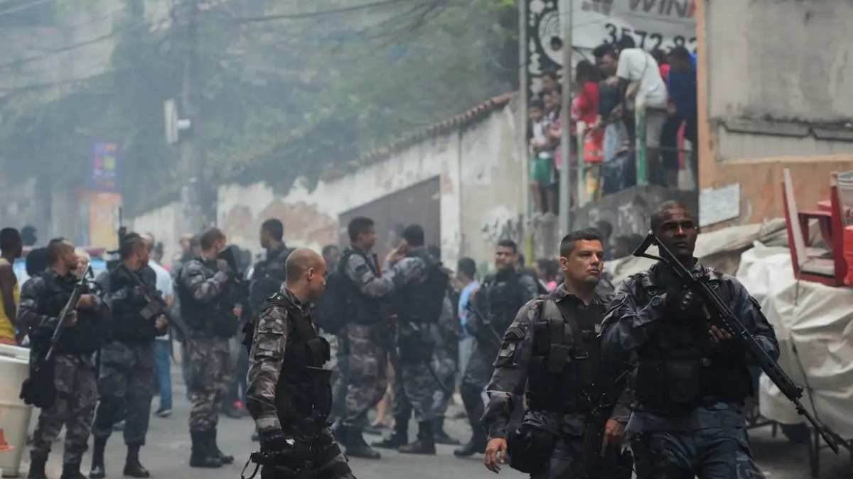 "Policiais fortemente armados em operação em comunidade, enquanto moradores observam das casas.