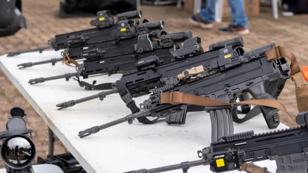 Fuzis modernos alinhados sobre uma mesa branca, equipados com miras ópticas e acessórios táticos, prontos para distribuição às forças de segurança.

