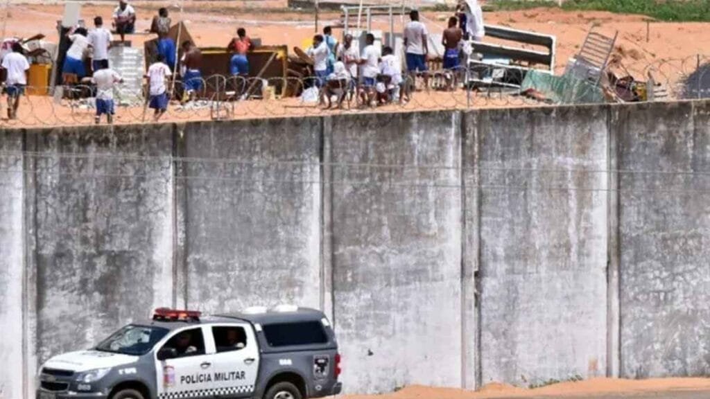 Presos aglomerados no topo do muro da penitenciária com objetos e barricadas improvisadas, enquanto viatura da Polícia Militar está posicionada do lado de fora