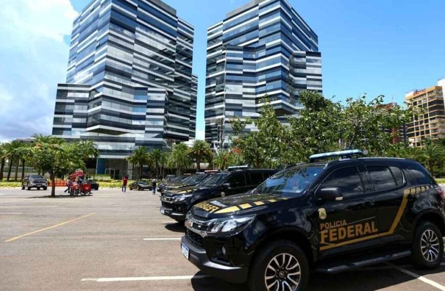 A imagem mostra viaturas da **Polícia Federal** estacionadas em frente a um conjunto de prédios modernos, de fachada espelhada, em área urbana.