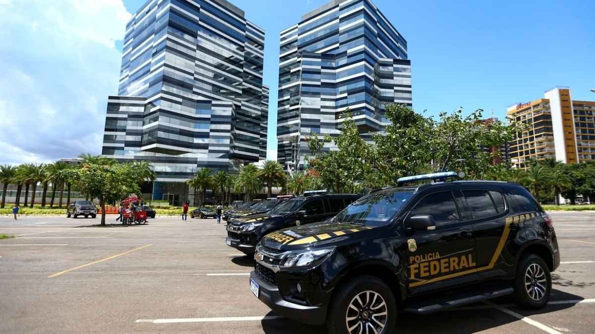 A imagem mostra viaturas da **Polícia Federal** estacionadas em frente a um conjunto de prédios modernos, de fachada espelhada, em área urbana.