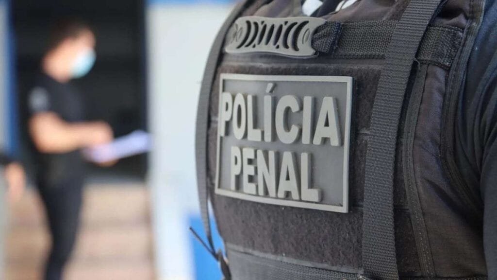 Colete tático com identificação “Polícia Penal” em destaque; ao fundo, pessoa desfocada usando máscara.
