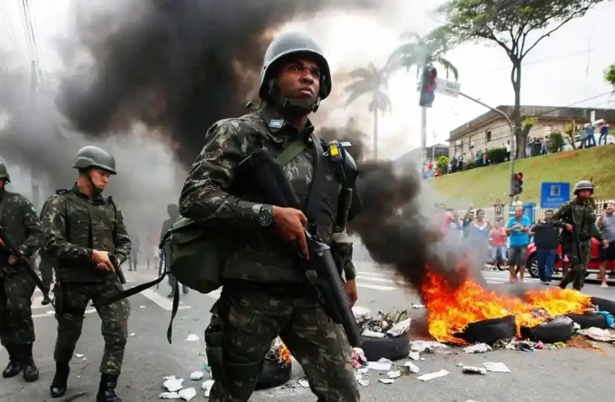 Espírito Santo, 2017: sem PM, a sociedade virou um caos - O ChatGPT disse: Soldados das Forças Armadas patrulham uma rua durante a crise de segurança no Espírito Santo, em 2017. À frente, um militar caminha armado enquanto, ao fundo, pneus e entulhos queimam.