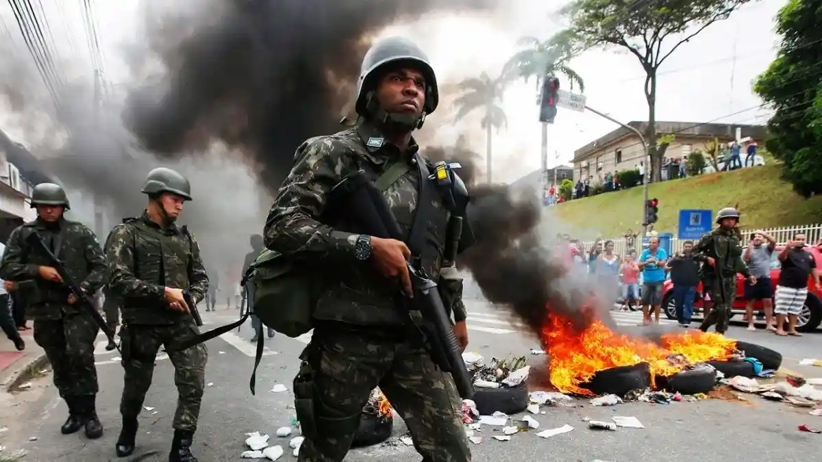 Espírito Santo, 2017: sem PM, a sociedade virou um caos - O ChatGPT disse: Soldados das Forças Armadas patrulham uma rua durante a crise de segurança no Espírito Santo, em 2017. À frente, um militar caminha armado enquanto, ao fundo, pneus e entulhos queimam.
