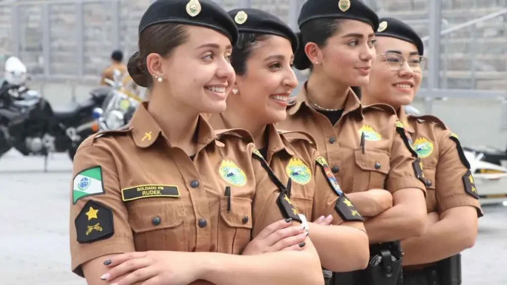 Quatro policiais militares femininas em uniforme marrom posam sorridentes lado a lado.