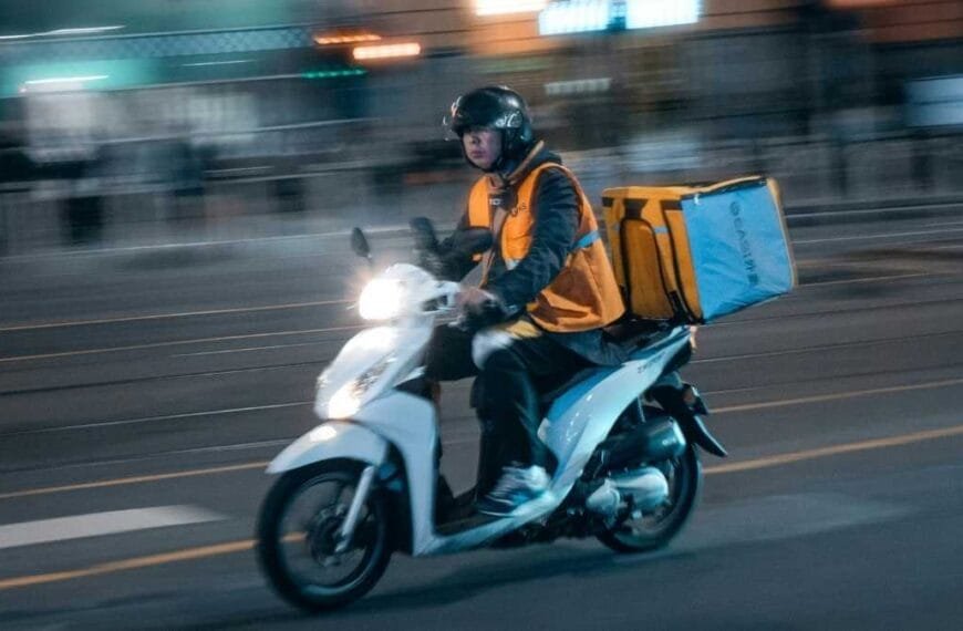 Entregador de moto circulando à noite com mochila térmica amarela nas costas e colete de segurança laranja.