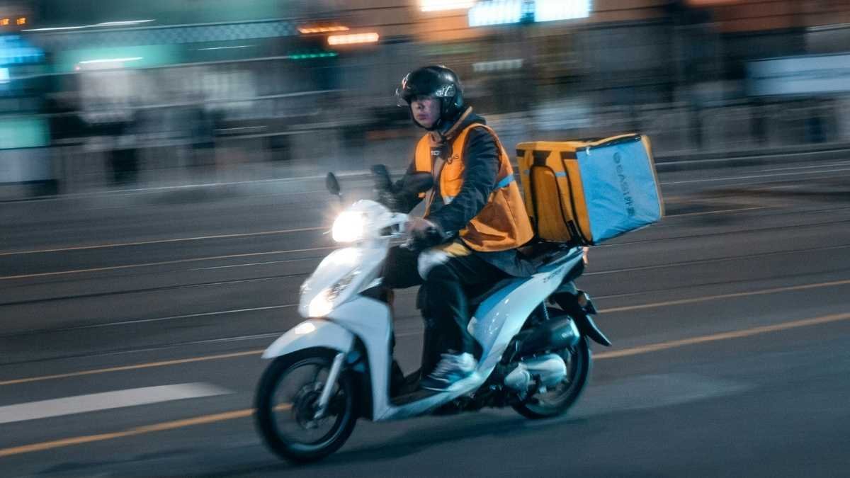 Entregador de moto circulando à noite com mochila térmica amarela nas costas e colete de segurança laranja.