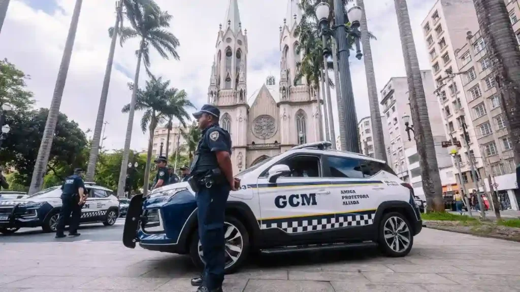 Guardas civis municipais ao lado de viaturas da GCM em praça pública, com a Catedral da Sé ao fundo.