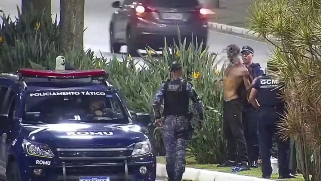 A imagem mostra uma ocorrência envolvendo a **Guarda Civil Municipal (GCM)**. Dois agentes abordam um homem sem camisa próximo a arbustos, enquanto outro guarda se aproxima. À frente, há uma viatura azul da **ROMU** com identificação de “Policiamento Tático”. A cena ocorre em via urbana, durante a noite.