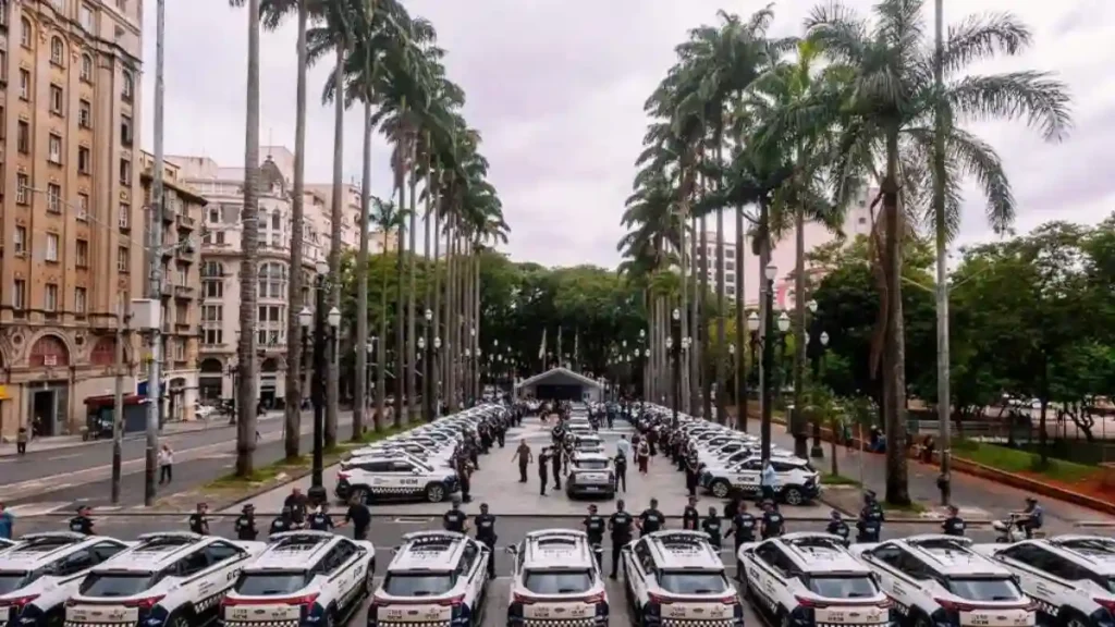 Fileira de viaturas da Guarda Civil Metropolitana de São Paulo estacionadas lado a lado em avenida com palmeiras, durante cerimônia oficial com agentes enfileirados.