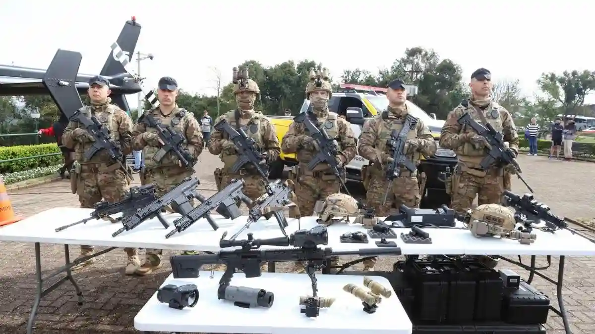 Fuzis israelenses: Paraná reforça polícias.Soldados das forças especiais do Paraná, em uniforme camuflado e armados com fuzis modernos, posam lado a lado atrás de mesas que exibem armas, capacetes e equipamentos táticos; ao fundo há viaturas e um helicóptero da segurança pública.