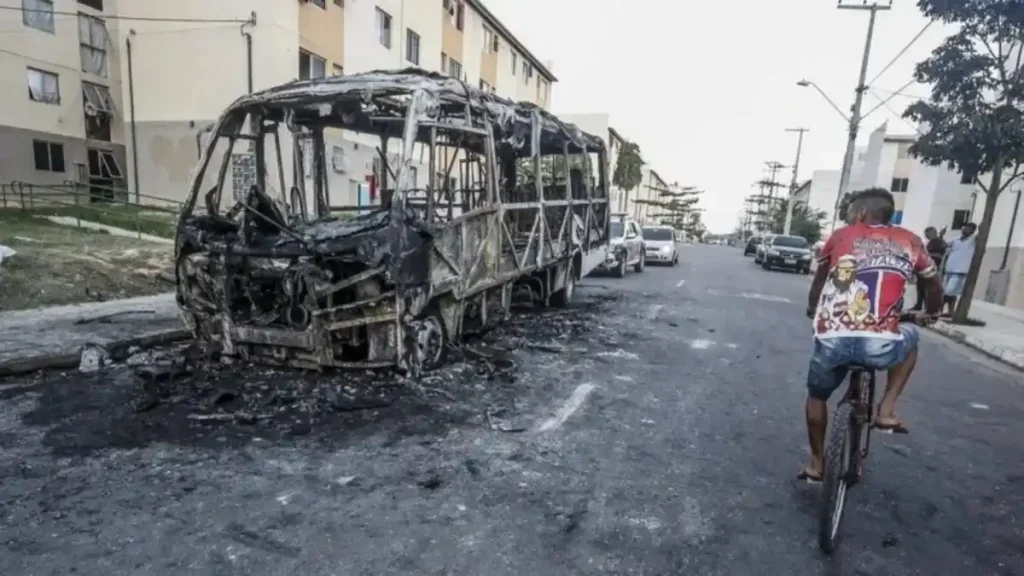 A imagem mostra um ônibus completamente queimado no meio de uma rua residencial. Em primeiro plano, um homem anda de bicicleta; ao fundo, moradores observam a cena. O cenário sugere ataque criminoso ou ação de facções.