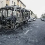 A imagem mostra um ônibus completamente queimado no meio de uma rua residencial. Em primeiro plano, um homem anda de bicicleta; ao fundo, moradores observam a cena. O cenário sugere ataque criminoso ou ação de facções.