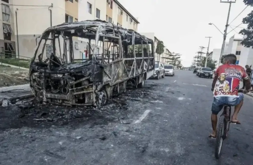 A imagem mostra um ônibus completamente queimado no meio de uma rua residencial. Em primeiro plano, um homem anda de bicicleta; ao fundo, moradores observam a cena. O cenário sugere ataque criminoso ou ação de facções.