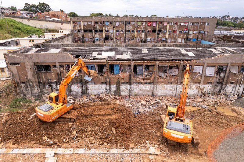 Duas escavadeiras laranjas trabalham na demolição de um prédio em condições precárias, com janelas quebradas, paredes deterioradas e roupas penduradas. Ao fundo, vê-se outro edifício também degradado, ocupado por moradores. O cenário transmite situação de vulnerabilidade habitacional.