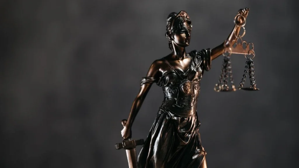 A imagem mostra uma estátua de bronze da deusa da Justiça, com os olhos vendados, segurando uma balança em uma das mãos e uma espada na outra.
