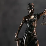 A imagem mostra uma estátua de bronze da deusa da Justiça, com os olhos vendados, segurando uma balança em uma das mãos e uma espada na outra.
