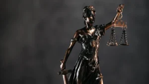 A imagem mostra uma estátua de bronze da deusa da Justiça, com os olhos vendados, segurando uma balança em uma das mãos e uma espada na outra.