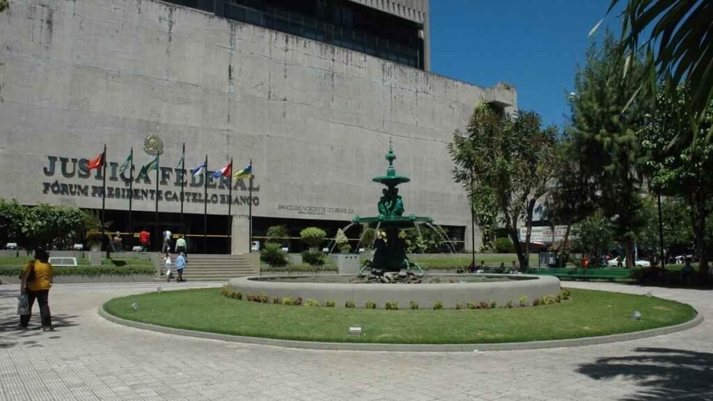 Prédio da Justiça Federal do Ceará, Fórum Presidente Castelo Branco, com fonte verde no jardim frontal e bandeiras hasteadas na entrada. Algumas pessoas caminham pelo local.