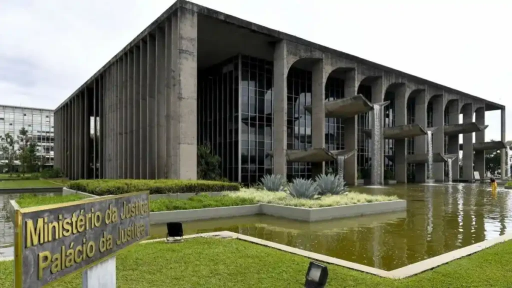 Fachada do Ministério da Justiça e Segurança Pública, em Brasília, com espelhos d’água e quedas artificiais em frente ao prédio. Uma placa indica “Ministério da Justiça – Palácio da Justiça”.