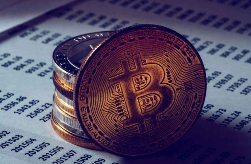 Operação EBDOX-Moeda física com símbolo do Bitcoin em destaque, apoiada sobre uma pilha de moedas, em cima de folha com números e dados financeiros impressos.