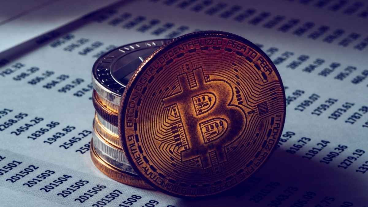 Operação EBDOX-Moeda física com símbolo do Bitcoin em destaque, apoiada sobre uma pilha de moedas, em cima de folha com números e dados financeiros impressos.