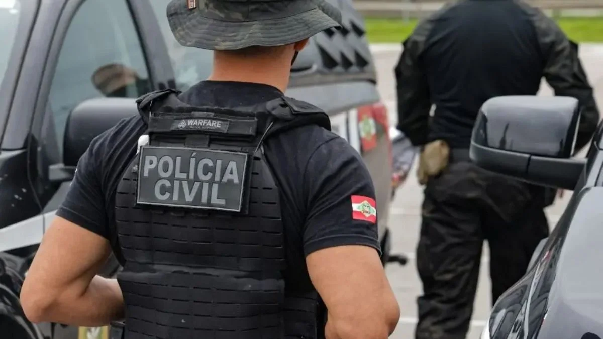 Por que o Brasil resiste ao ciclo completo de polícia? Policial civil de colete tático, visto de costas, com identificação “Polícia Civil” e bandeira de Santa Catarina no uniforme, em operação junto a viaturas.