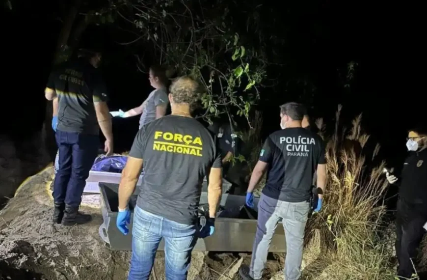 Icaraíma/PR: corpos de desaparecidos são encontrados-Na imagem, agentes da Força Nacional e da Polícia Civil do Paraná trabalham em uma área de mata durante a noite. Eles utilizam luvas e máscaras de proteção enquanto manuseiam uma estrutura metálica, ao lado de um corpo envolto em plástico.