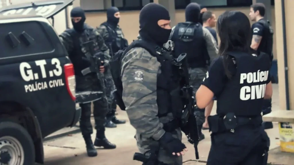 Grupo de policiais civis fortemente armados, alguns com balaclavas e fardas camufladas, ao lado de viatura identificada como G.T.3; em primeiro plano, policial civil conversa com agente tático durante operação.