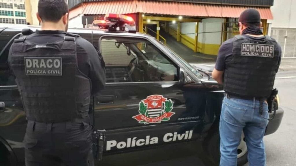 Narcoestado instalado: PCC executa ex-Delegado Geral-Dois policiais civis aparecem de costas ao lado de uma viatura preta da Polícia Civil de São Paulo, identificada com o brasão do estado. Um deles veste colete com a inscrição “DRACO” e o outro “Homicídios”, indicando divisões especializadas. A cena ocorre em área urbana, em frente a um estacionamento coberto.