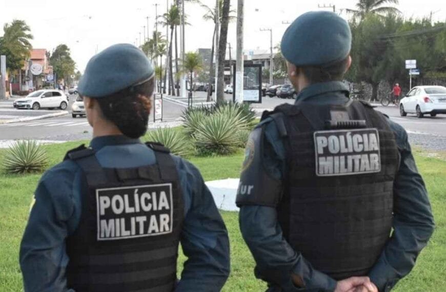Dois policiais militares, de costas, observam uma via urbana movimentada durante patrulhamento. Ambos usam uniforme azul e colete balístico com a inscrição "POLÍCIA MILITAR".