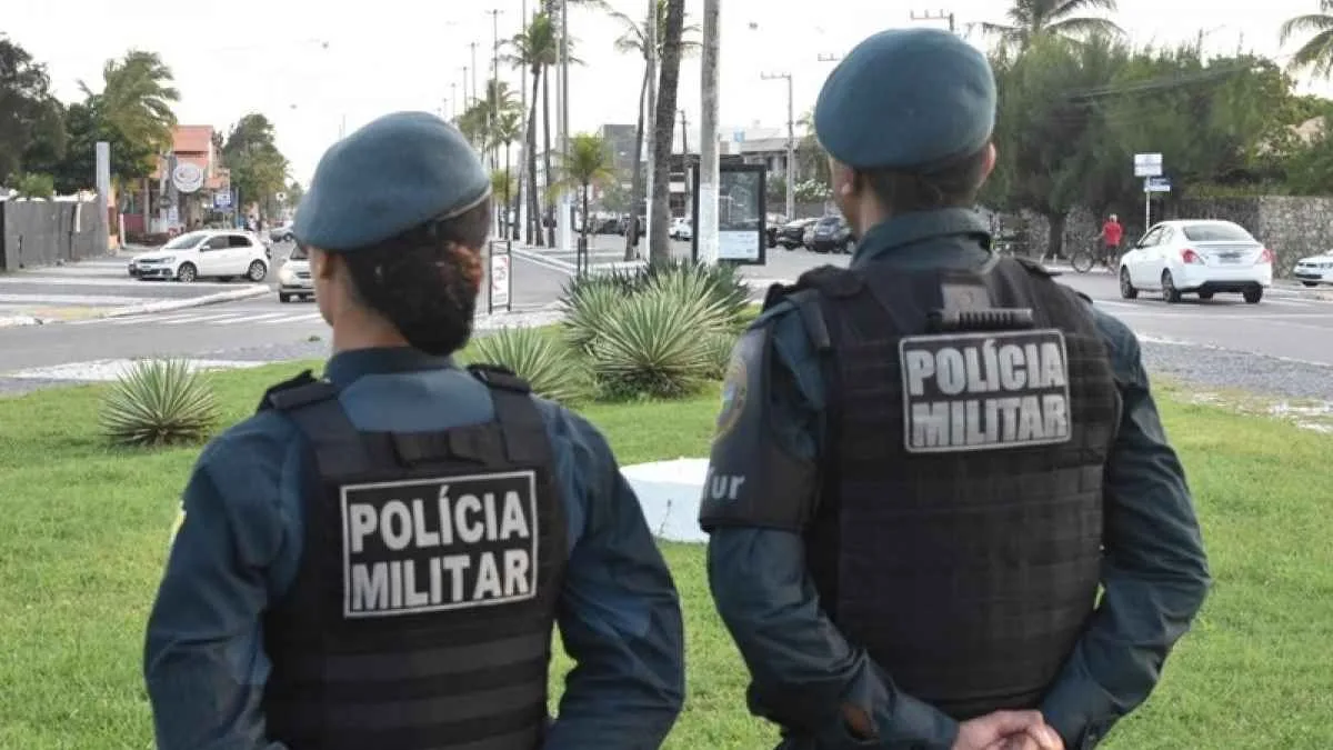 Dois policiais militares, de costas, observam uma via urbana movimentada durante patrulhamento. Ambos usam uniforme azul e colete balístico com a inscrição "POLÍCIA MILITAR".