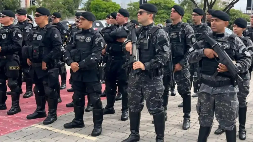 Emenda à PEC 18 cria ciclo completo de polícia-A imagem mostra um grupo de policiais militares fortemente armados e equipados, usando uniformes táticos pretos e cinza camuflado, boinas pretas e óculos escuros. Eles estão perfilados em formação ao ar livre, aparentando estar em momento de instrução, solenidade ou operação.