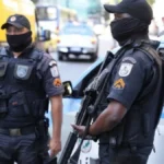 41 PMs mortos - Dois policiais militares do Rio de Janeiro aparecem em serviço, usando coletes táticos, máscaras e armamento, ao lado de uma viatura da corporação estacionada em via urbana. aposentadoria especial
