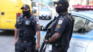 41 PMs mortos - Dois policiais militares do Rio de Janeiro aparecem em serviço, usando coletes táticos, máscaras e armamento, ao lado de uma viatura da corporação estacionada em via urbana. aposentadoria especial