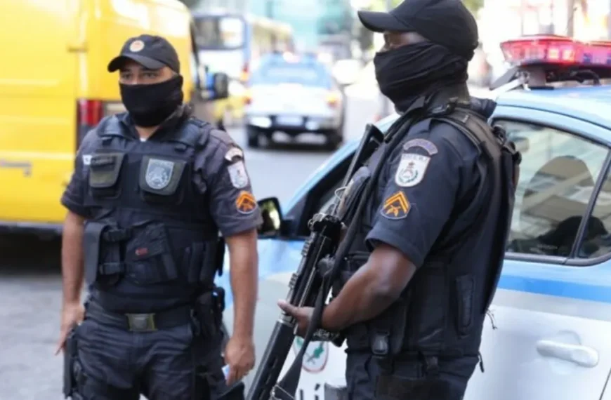 41 PMs mortos - Dois policiais militares do Rio de Janeiro aparecem em serviço, usando coletes táticos, máscaras e armamento, ao lado de uma viatura da corporação estacionada em via urbana.