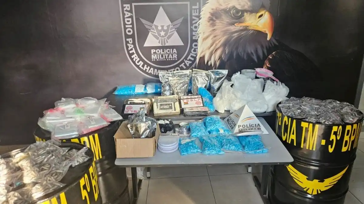 Imóvel alugado por idosa vira laboratório de drogas - Mesa exibe grande quantidade de drogas, insumos químicos e embalagens apreendidas pela Polícia Militar de Minas Gerais; ao fundo, banner do Batalhão com logotipo e símbolo de águia.