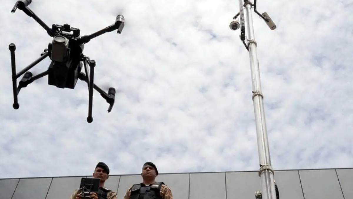 Drones: de recreação a uso em perícia criminal-Dois policiais militares observam o voo de um drone de monitoramento, próximo a um poste equipado com câmeras de vigilância, sob um céu parcialmente nublado.