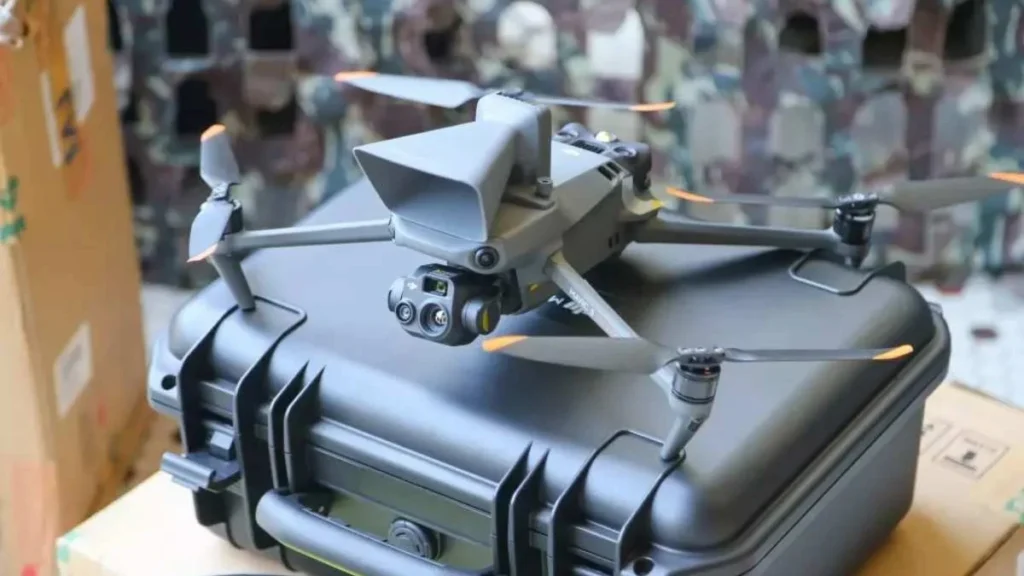 Drone de vigilância pousado sobre um case de transporte rígido, equipado com câmeras e sensores, em ambiente com fundo camuflado.