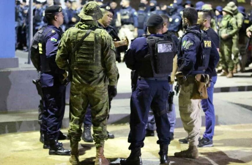 Grupo de policiais de diferentes corporações, incluindo militares e rodoviários federais, reunidos em operação noturna, usando fardas e coletes táticos, em pé em área iluminada.