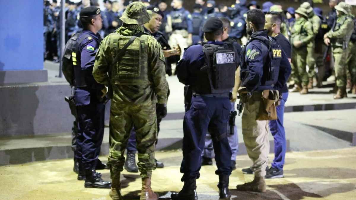 Grupo de policiais de diferentes corporações, incluindo militares e rodoviários federais, reunidos em operação noturna, usando fardas e coletes táticos, em pé em área iluminada.