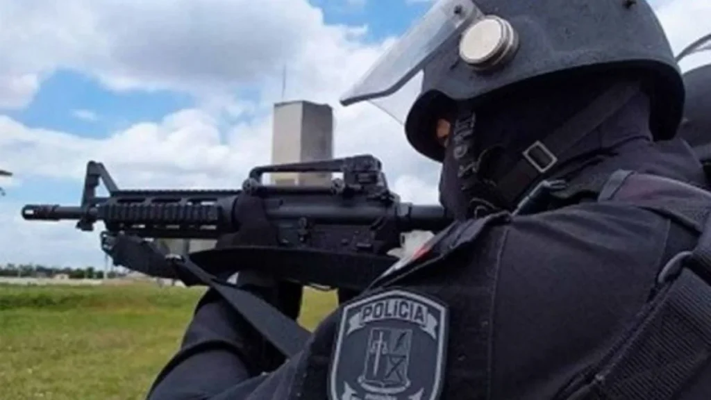 Policial penal fortemente equipado mira com fuzil durante operação de segurança.